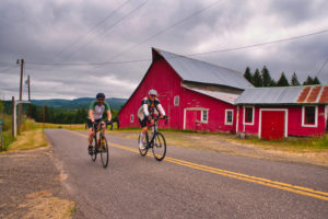 2 bicyclists riding in front of a big red barn